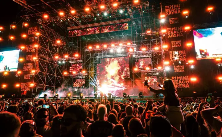 Folla in delirio al Rimini Beach Arena, luci psichedeliche e musica pulsante per un evento indimenticabile.