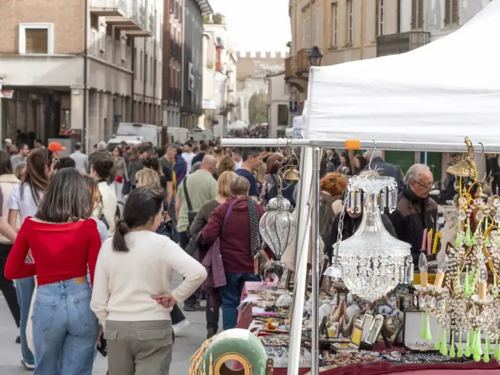 Folla animata in un mercatino usato a Rimini, tra bancarelle con lampadari di cristallo e oggetti d'antiquariato.