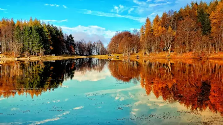 Lago specchiato con alberi autunnali e verdi, ideale per il trekking in Emilia Romagna. Cielo azzurro con nuvole bianche.